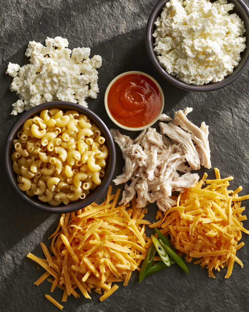 Flat lay of buffalo chicken mac and cheese ingredients: macaroni, cottage cheese, chicken, buffalo sauce, cheddar