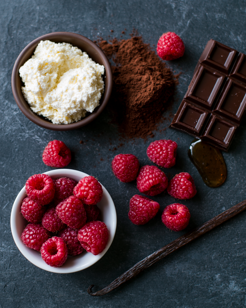 Flat lay of dark chocolate raspberry mousse ingredients: cottage cheese, cocoa, raspberries, dark chocolate, honey, vanilla