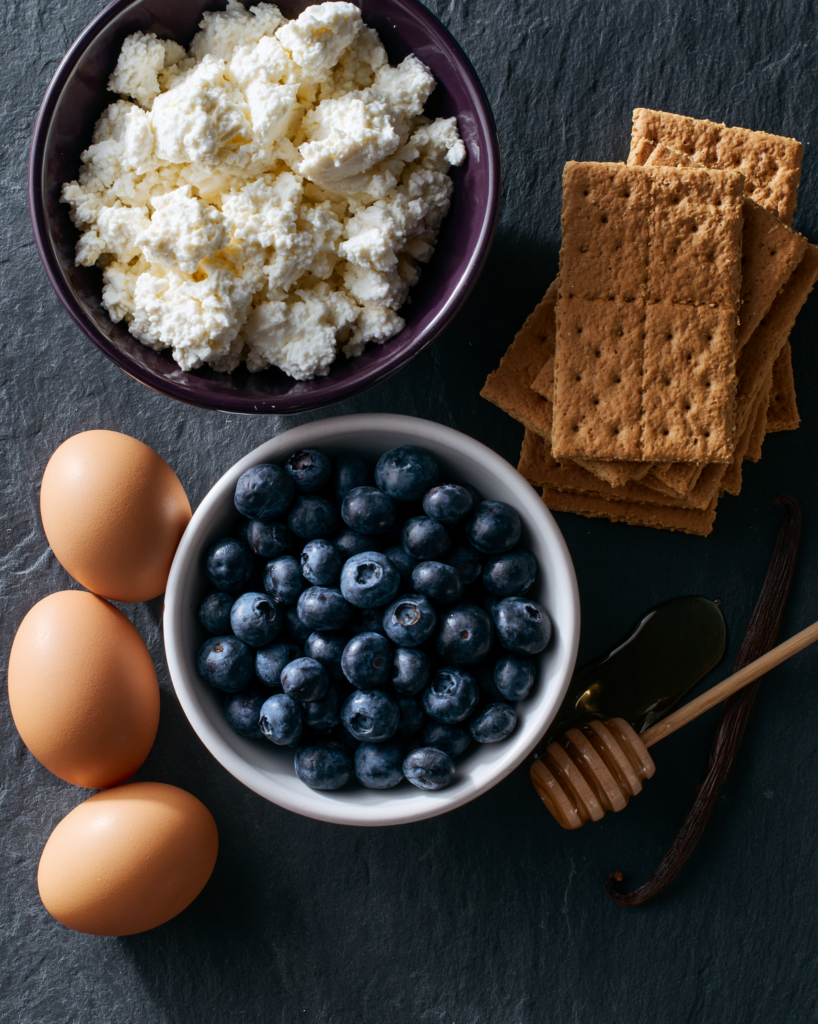 Flat lay of blueberry cheesecake bars ingredients: cottage cheese, blueberries, graham crackers, eggs, honey