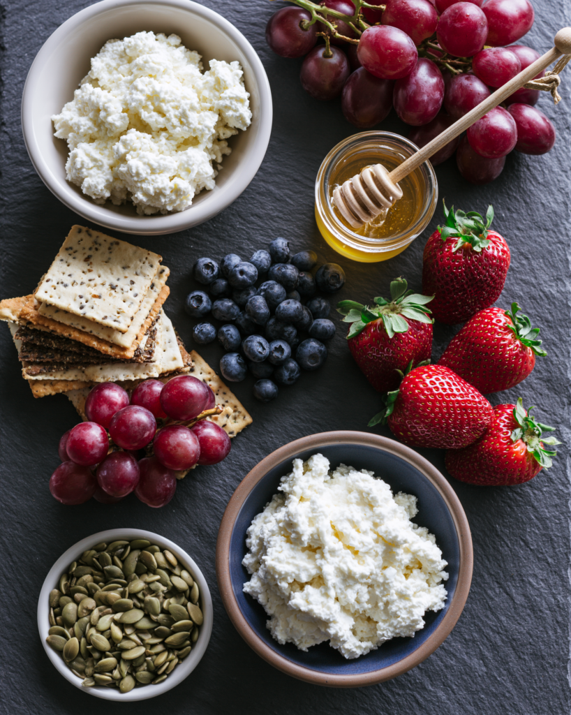 Flat lay of cottage cheese snack board components: cottage cheese, honey, crackers, berries, grapes, seeds