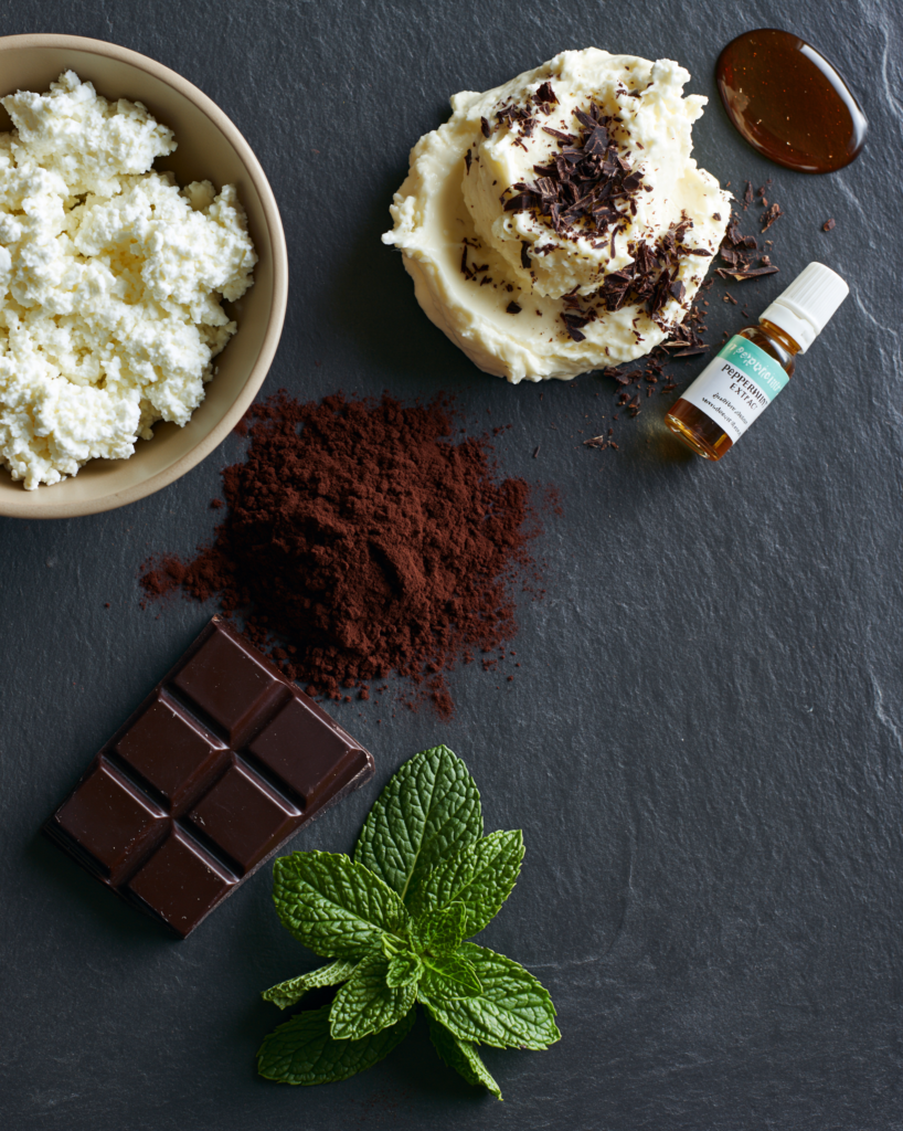 Flat lay of mint chocolate mousse ingredients: cottage cheese, cocoa, dark chocolate, fresh mint, peppermint extract